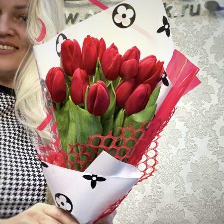 Букет из 15 красных тюльпанов в стильной упаковке с красной сеткой, украшенный декоративной бумагой с узорами, в руках девушки.